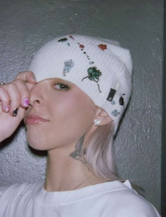 Eye-level portrait of a young woman wearing a decorated white beanie, white sweatshirt, and a butterfly tattoo on her neck in a gray room.