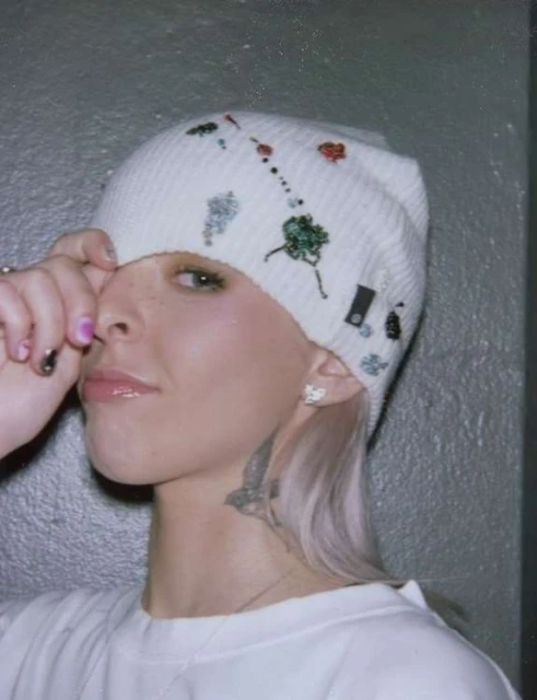 Eye-level portrait of a young woman wearing a decorated white beanie, white sweatshirt, and a butterfly tattoo on her neck in a gray room.