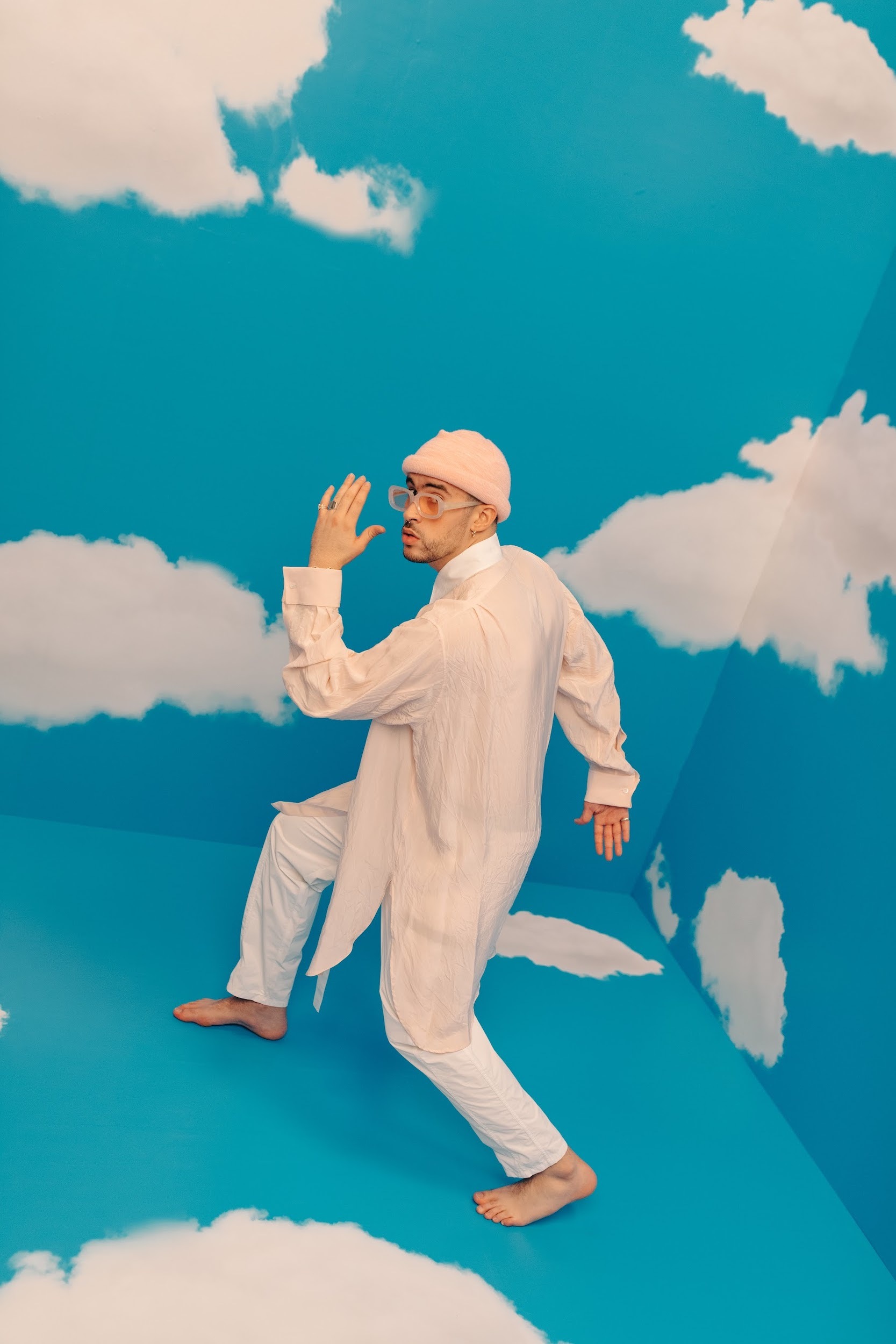 A man in a pink beanie, white shirt, and pants strikes a pose in a room with a blue wall and floor painted with clouds.