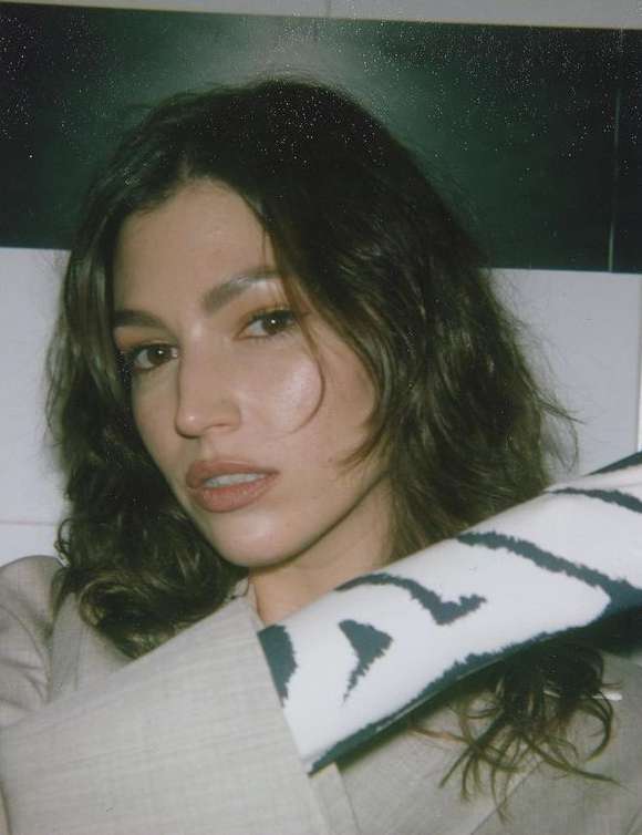 Close-up portrait of a woman with wavy brown hair and a beige blazer, holding a patterned fabric, against a dark and white background.