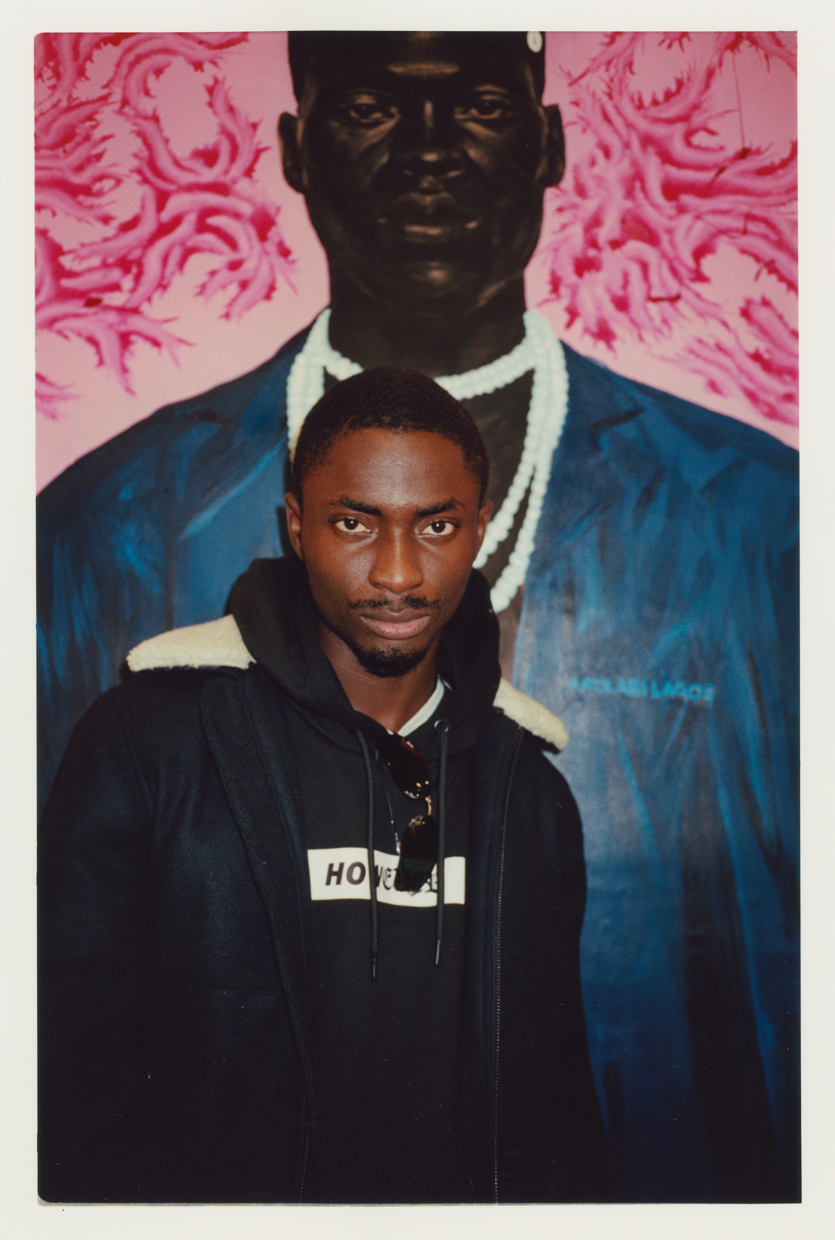 Portrait of an adult male in a hooded jacket, standing in front of a painting of a man with a pink floral background.