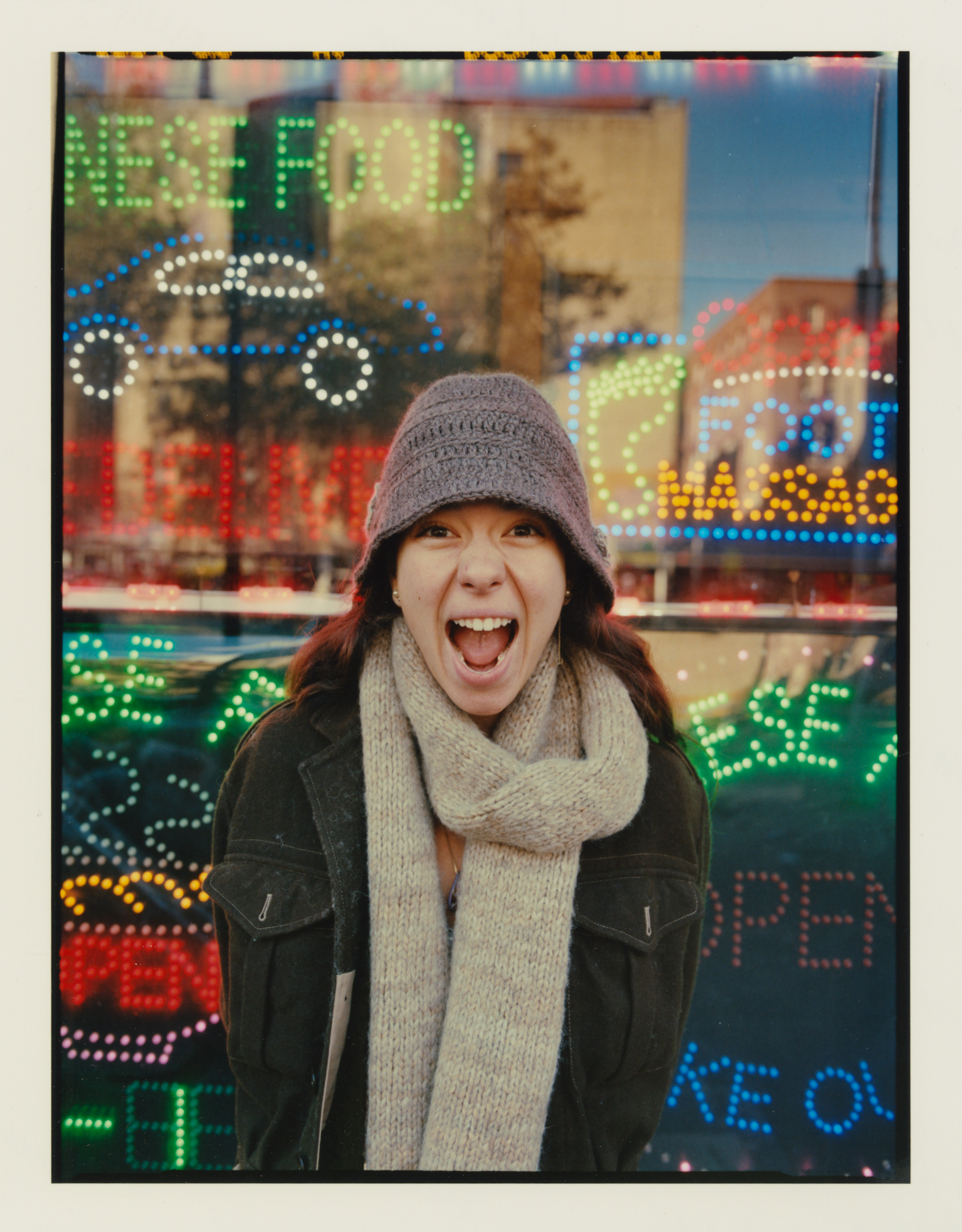 Expressive woman in hat and scarf shouts against colorful neon restaurant signage on city street, captured in a medium portrait shot.