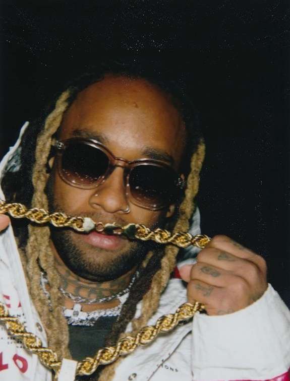 Close-up portrait of a man wearing dark sunglasses and holding a thick gold rope chain up to his mouth against a dark background.