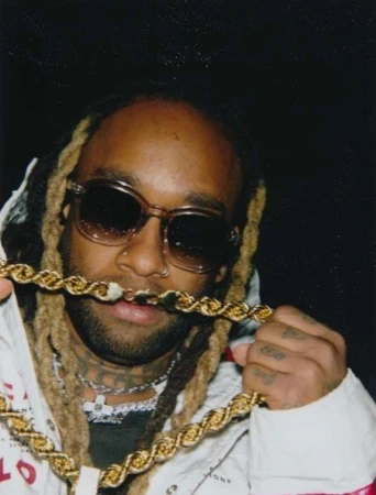 Close-up portrait of a man wearing dark sunglasses and holding a thick gold rope chain up to his mouth against a dark background.