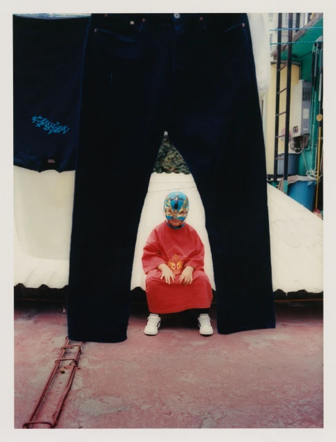 Child in wrestling mask sits in front of oversized jeans and t-shirt on a pink surface with a building in the background.