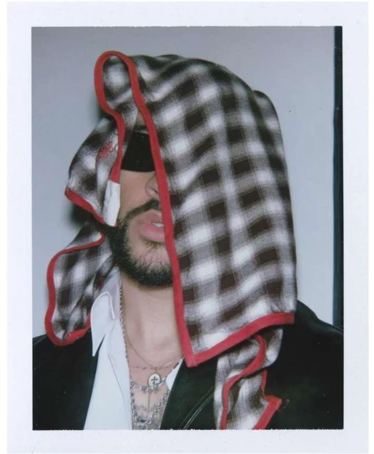 Portrait of a person wearing a black jacket and draped in a plaid textile covering their head, face, and hair, necklaces visible.