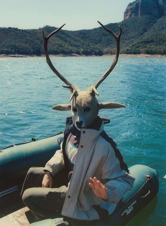 A person wearing a deer mask sits in a boat on a lake with mountains in the background, under bright daylight and sunny weather.