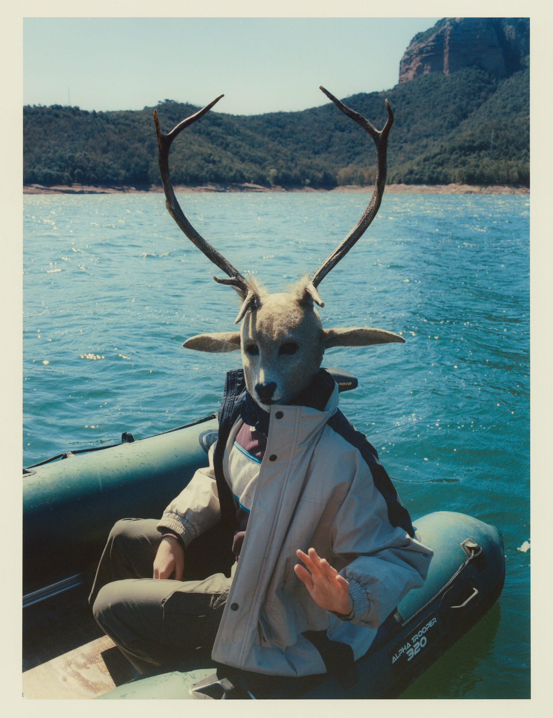 Person in a deer mask sits in a green inflatable boat on a lake with mountains in the background on a sunny day.