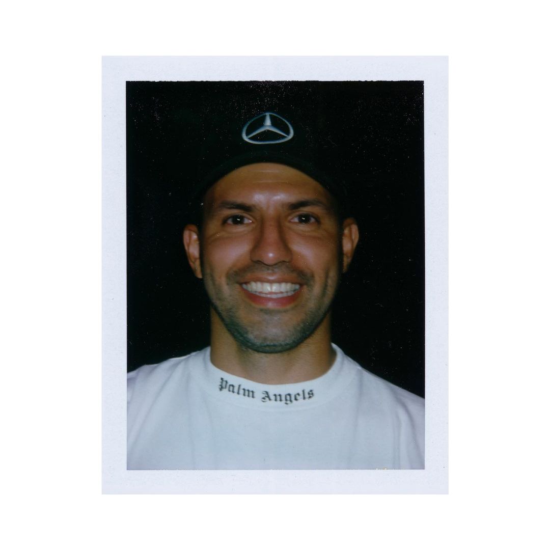 Close-up portrait of a smiling adult male wearing a Mercedes logo baseball cap and a Palm Angels t-shirt, set against a dark background.