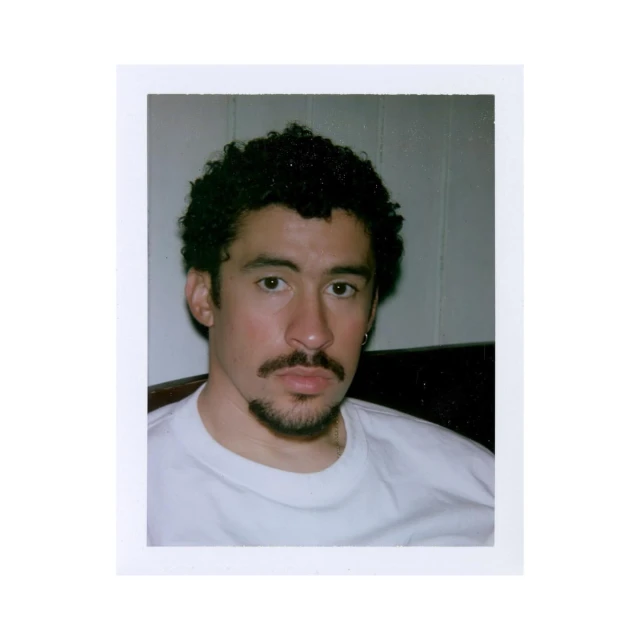 Close-up portrait of a man with dark, curly hair and a goatee, wearing a white t-shirt against a neutral, indoor background.