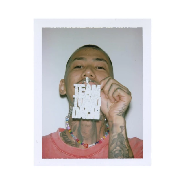 Close-up of a young adult wearing a colorful beaded necklace, tattoos, and holding a pendant with text on it against a white backdrop.