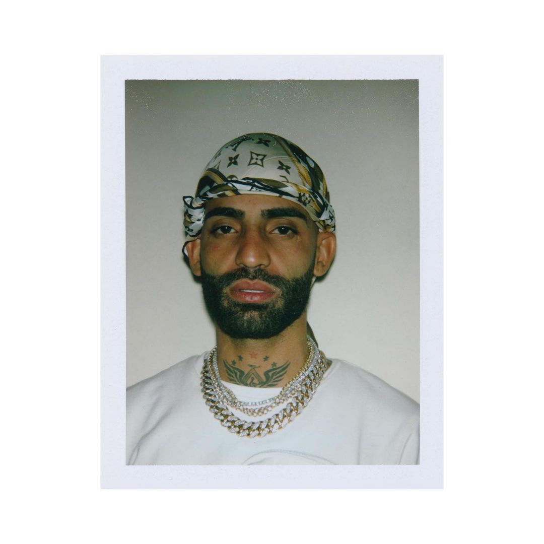 Close-up portrait of a bearded adult male wearing a patterned headscarf and several gold chain necklaces against a neutral background.