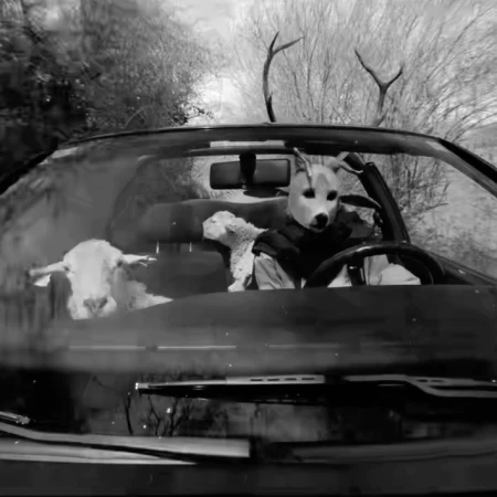 A person in a wolf mask drives a convertible car with two sheep visible in the passenger seat, in a black and white photograph.