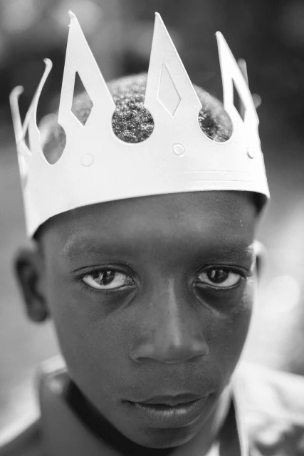 Black and white close-up of a young, dark-skinned boy wearing a white paper crown, looking directly at the camera with a serious expression.