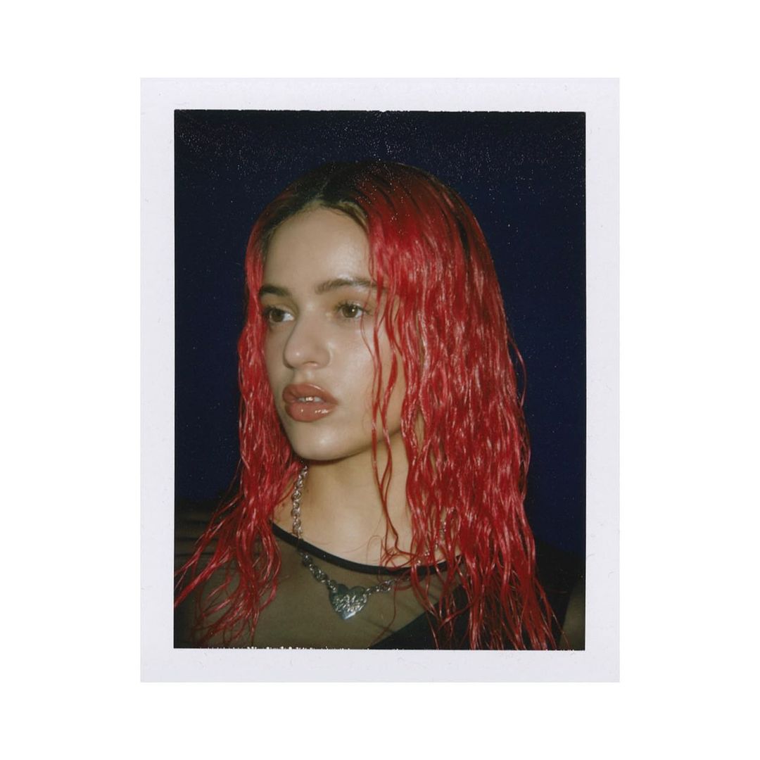 Close-up portrait of a young woman with red hair and a chain necklace, posing indoors with a neutral expression and dark background.