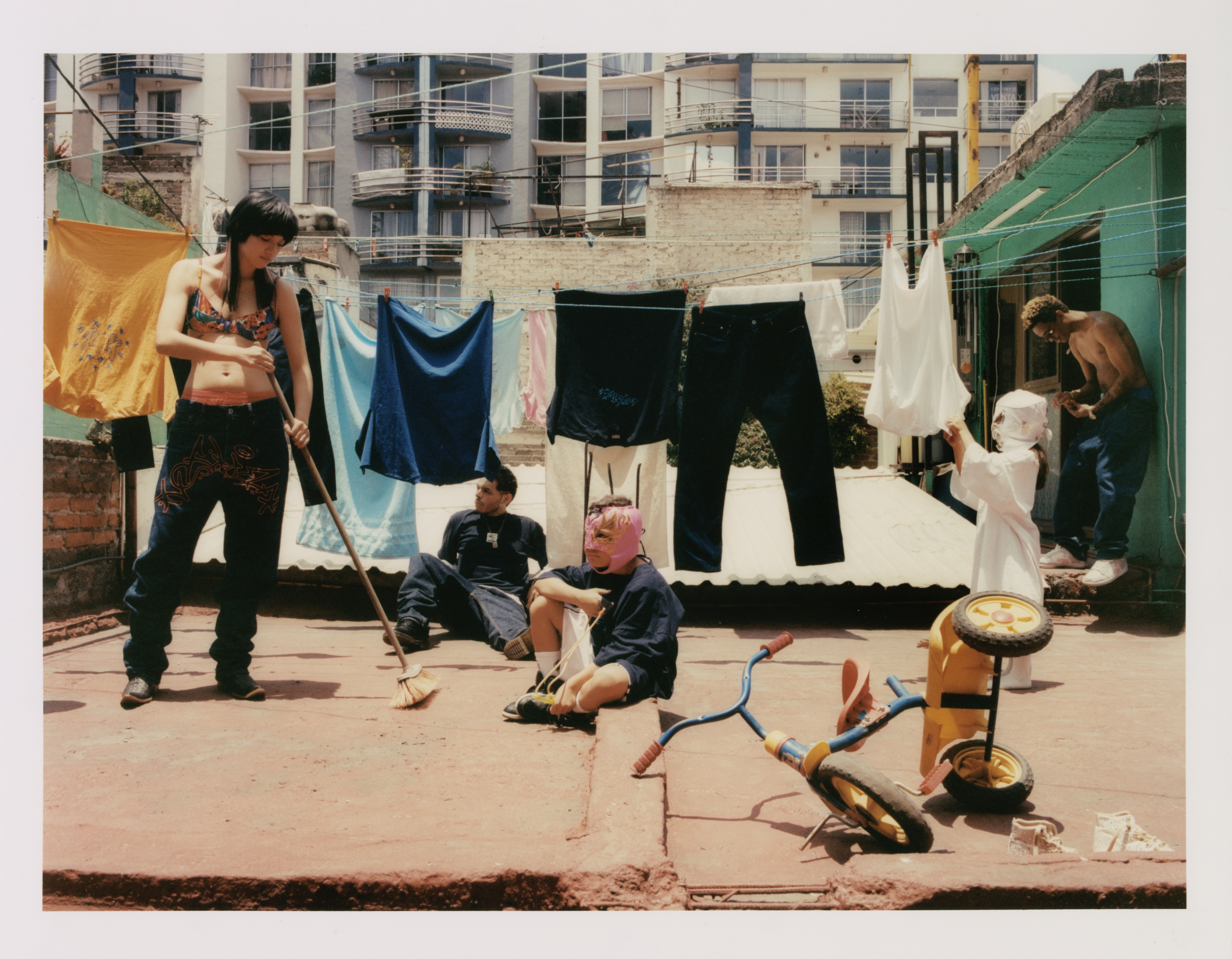 Outdoor scene on a rooftop with clothes hanging on a line, several people present, and a child's tricycle in the foreground.