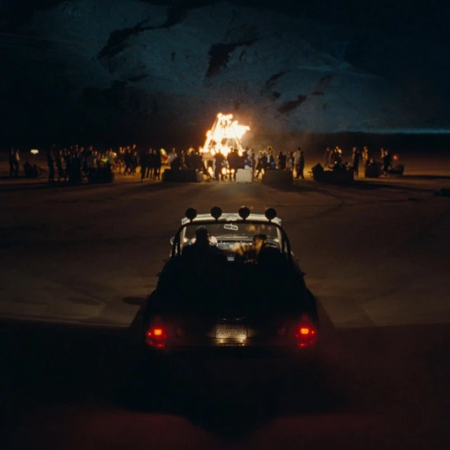 Night scene shows a group of people near a large bonfire, viewed from the rear of a car with headlights, set against mountains.