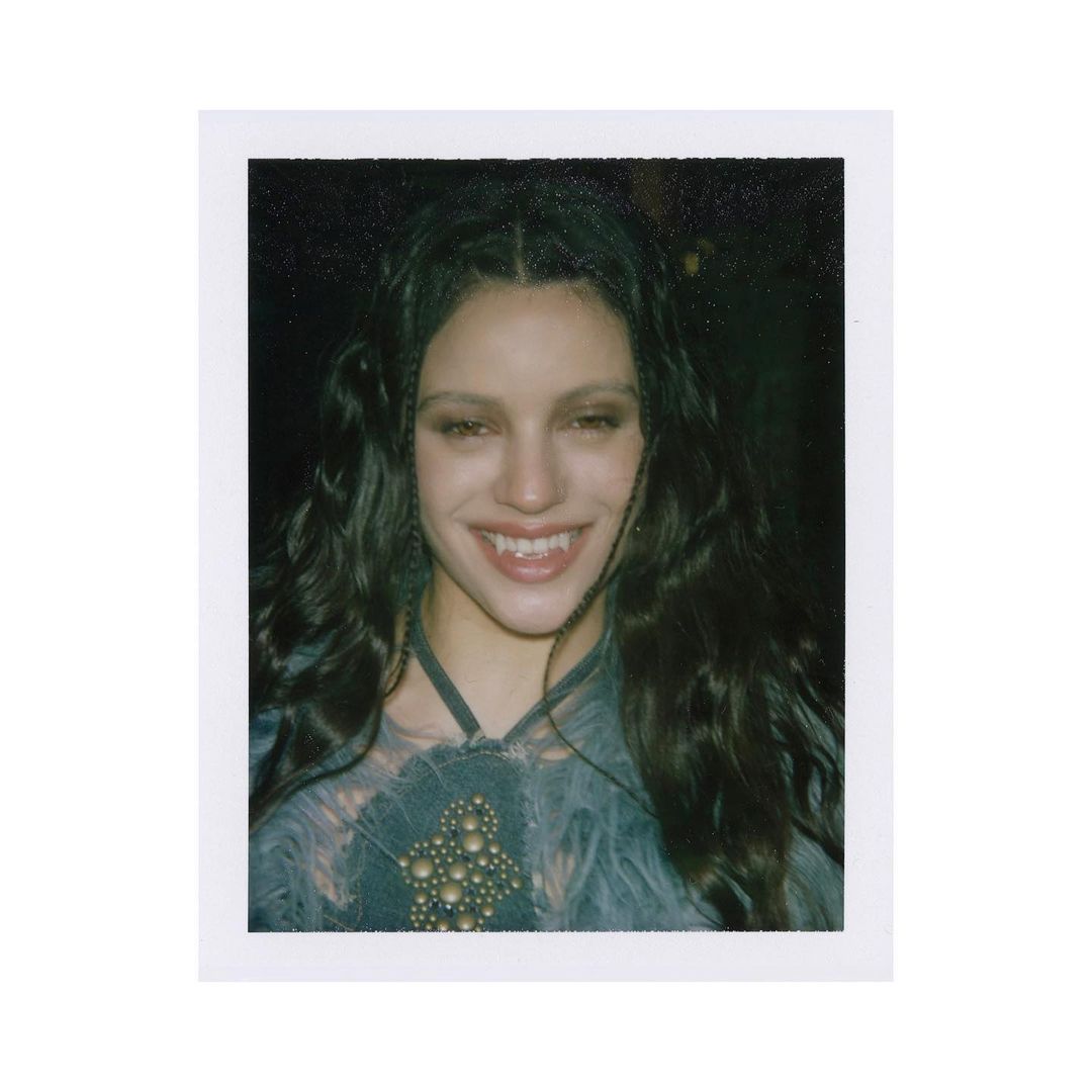 Close-up portrait of a smiling adult woman with long, wavy, dark hair, wearing an elaborate top, against a dark background.