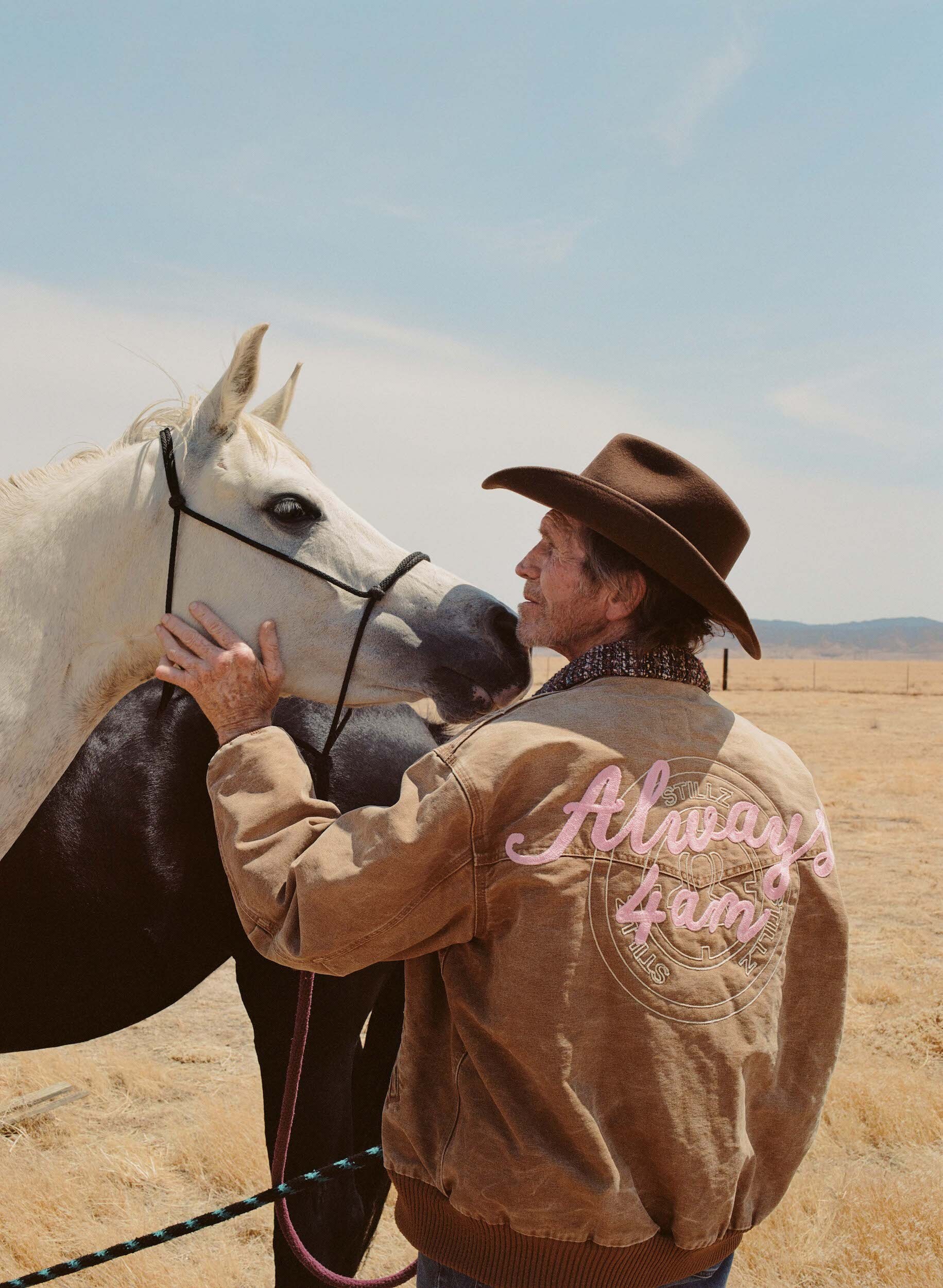 A man in a cowboy hat is petting a white horse in a sunny, rural landscape, wearing a jacket with the text 'Always 4am'.