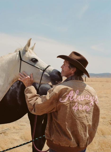 A man in a cowboy hat is petting a white horse in a sunny, rural landscape, wearing a jacket with the text 'Always 4am'.