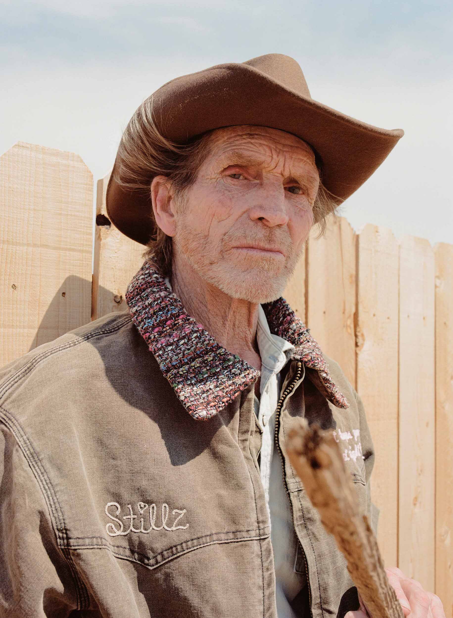 Portrait of an elderly man wearing a cowboy hat and jacket, standing outdoors with a wooden fence in the background on a sunny day.