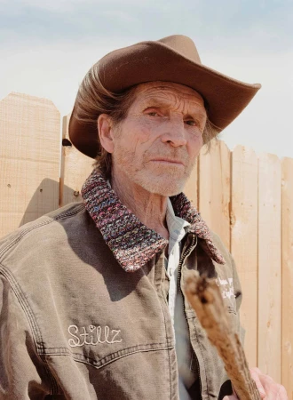 Portrait of an elderly man wearing a cowboy hat and jacket, standing outdoors with a wooden fence in the background on a sunny day.