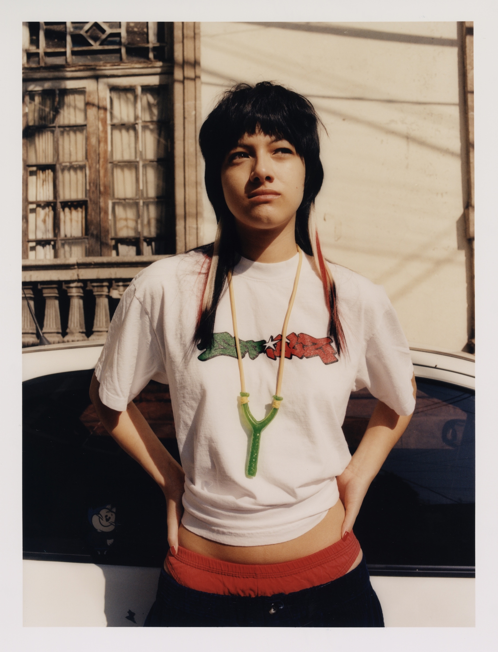 Portrait of a young woman with black hair and a graphic tee, posing outdoors with hands on hips against an urban setting and a white car.