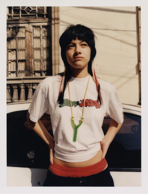 Portrait of a young woman with black hair and a graphic tee, posing outdoors with hands on hips against an urban setting and a white car.