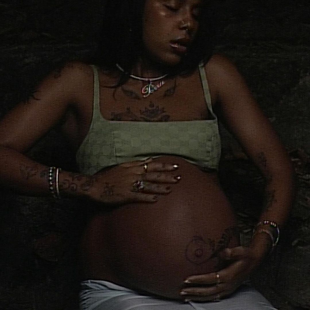 Pregnant woman with olive skin wearing a green top, cradling her baby bump, and adorned with jewelry in a dimly lit indoor setting.