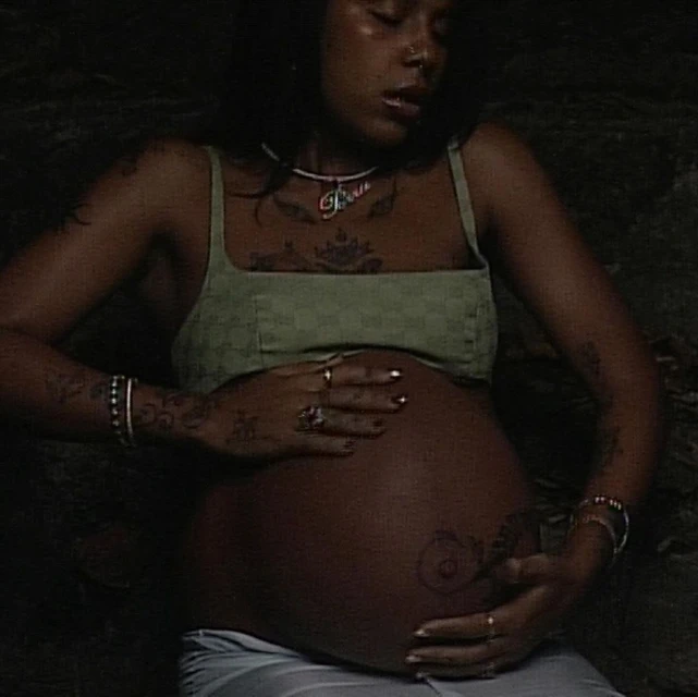 Pregnant woman with olive skin wearing a green top, cradling her baby bump, and adorned with jewelry in a dimly lit indoor setting.