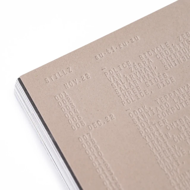 Close-up of a stack of paper with text embossed on the top sheet, featuring dates, titles, and locations in a textured, monochromatic composition.