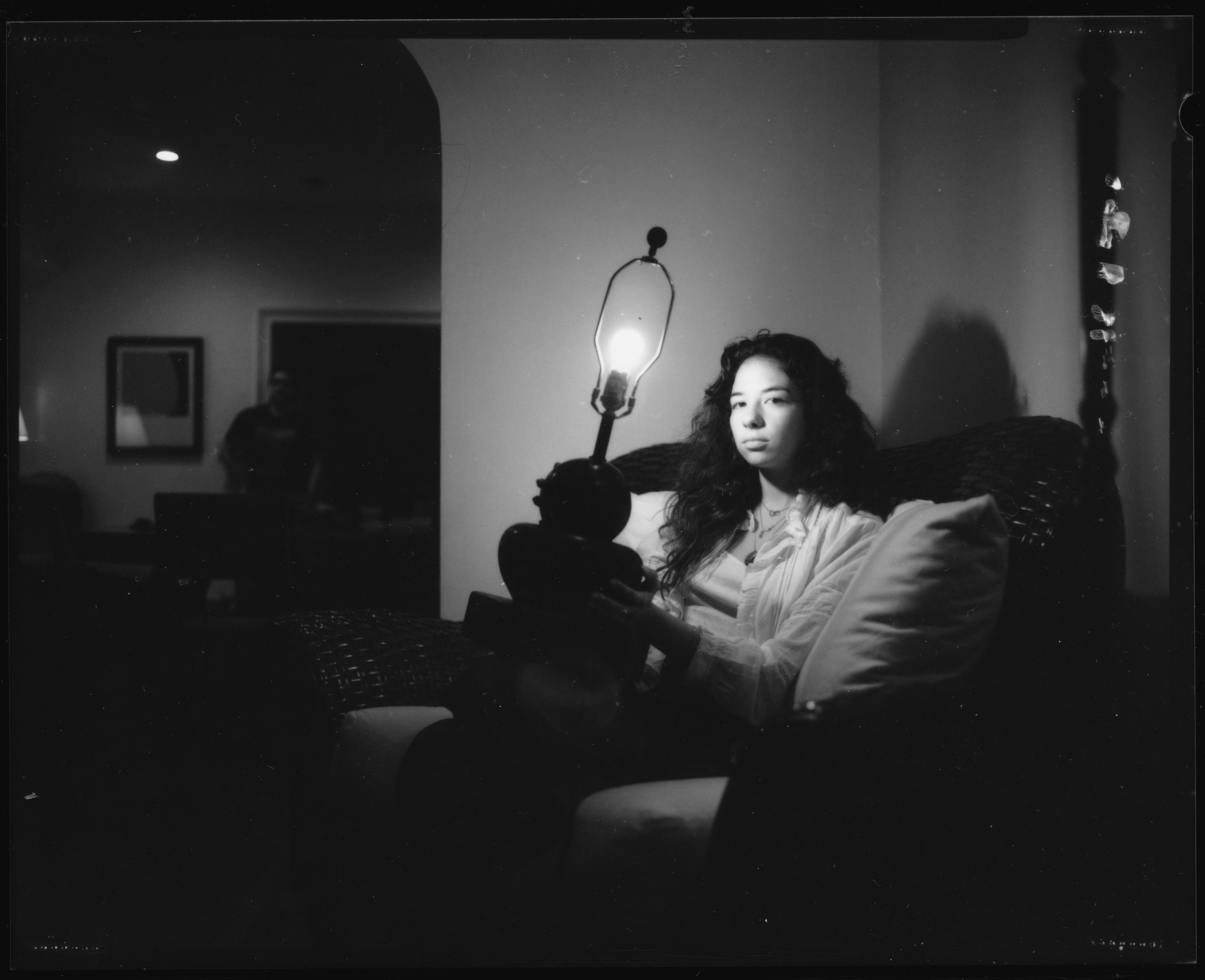 Monochrome portrait of a woman holding a lit table lamp while seated on a sofa in a dimly lit living room.