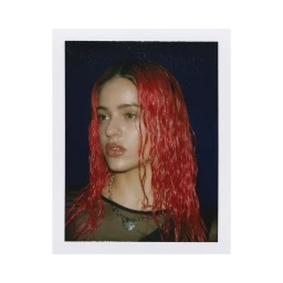 Polaroid photograph of singer Rosalia with wet red hair and a heart necklace against a dark background