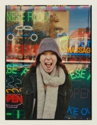 A woman wearing a grey knit bucket hat and beige scarf with her mouth open in a joyful expression in front of neon signs.