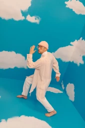 Man wearing a pink beanie and white tunic posing in a studio set with blue sky and cloud painted walls.