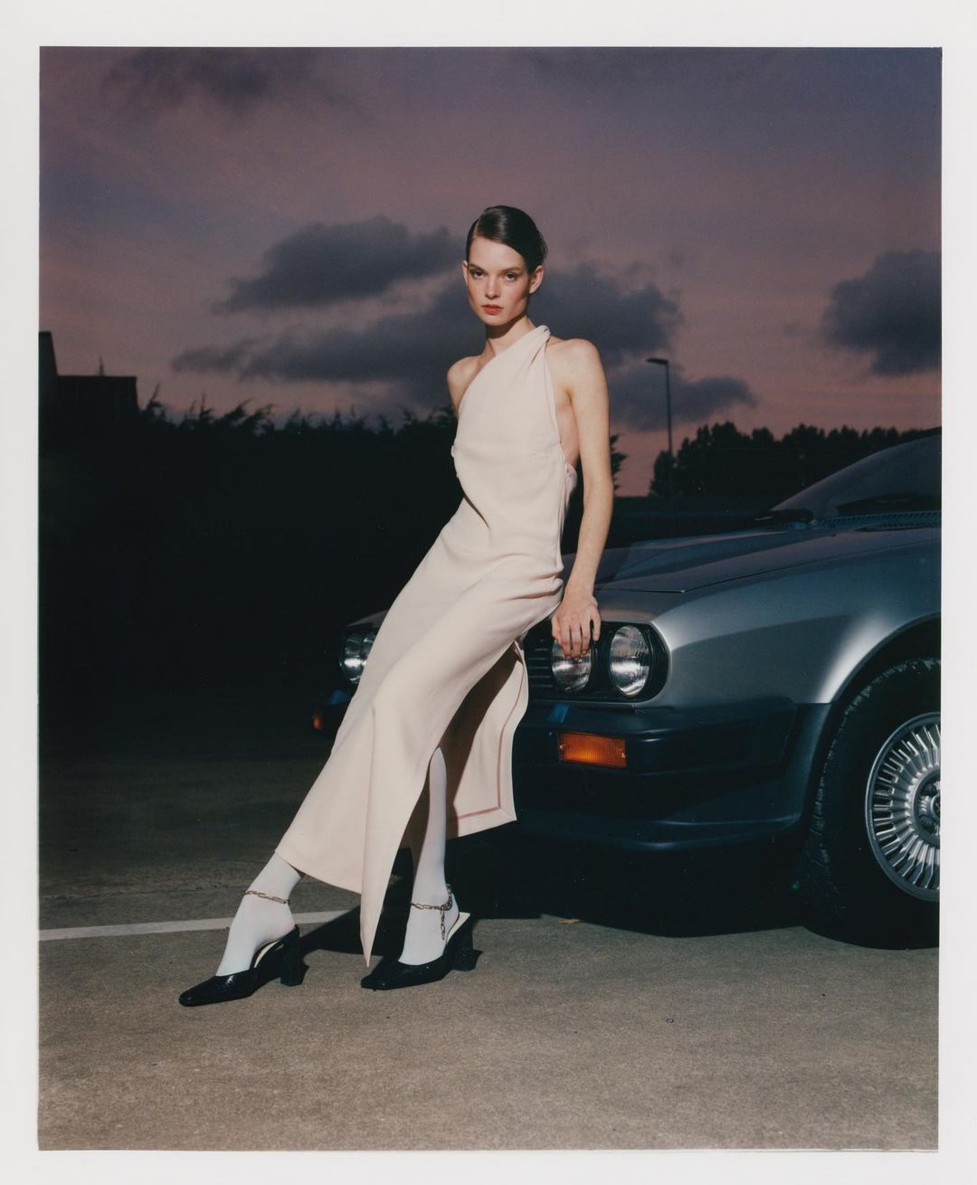 A female model in a light gown poses next to a classic silver car at dusk, creating a fashion-forward and stylized outdoor composition.