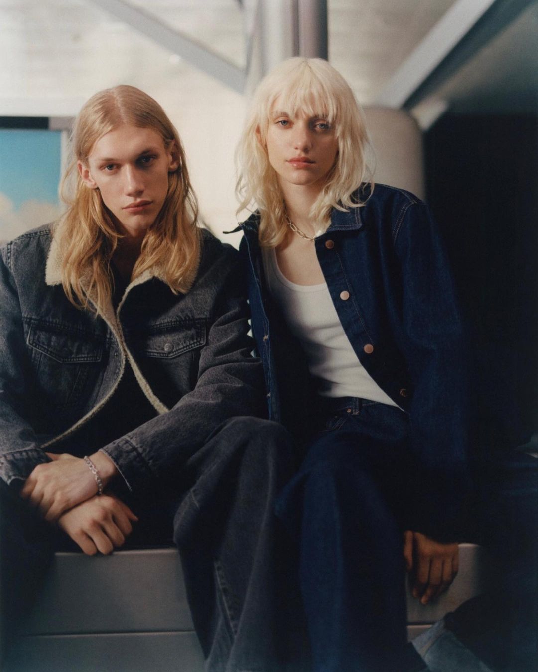 Two fashion models, an adult male and an adult female, pose indoors wearing denim jackets and pants, seated against a neutral background.