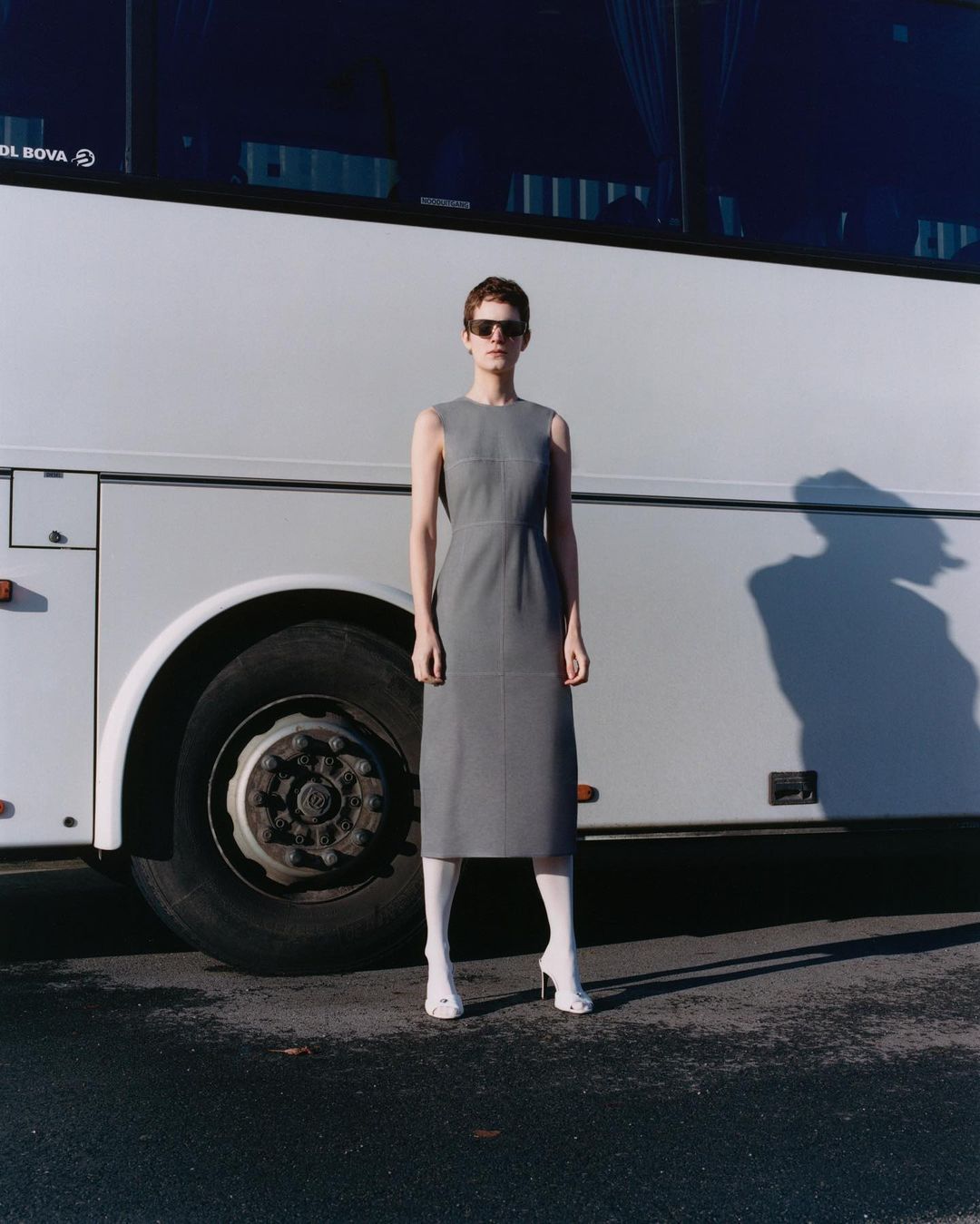 Woman in grey dress and white boots stands in front of a white bus, wearing sunglasses in an outdoor setting on a sunny day.
