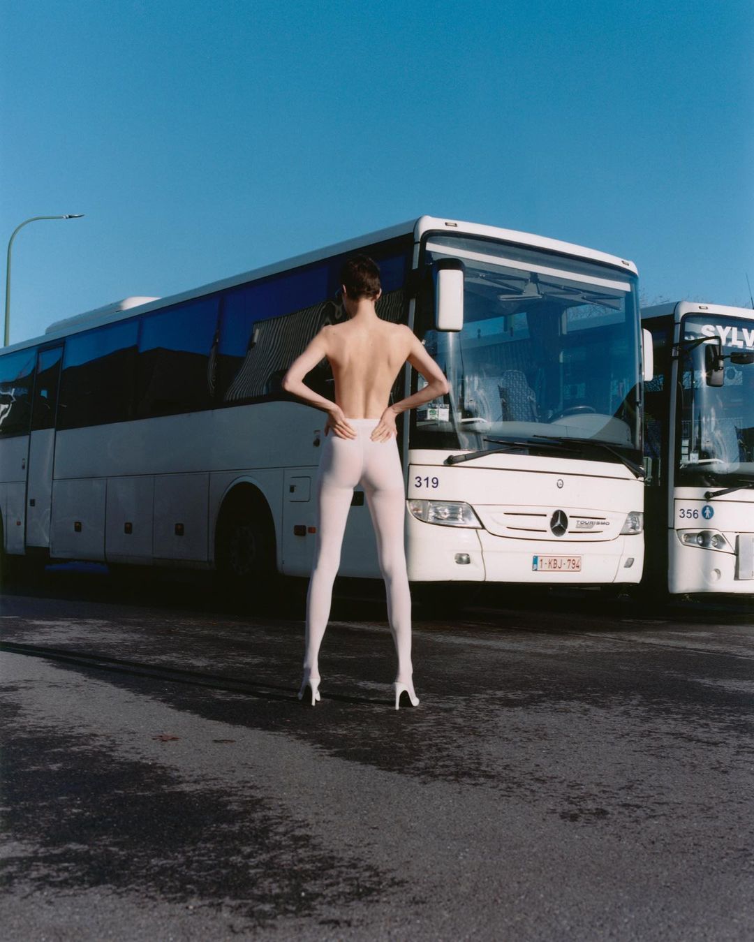 Back view of a slim woman in sheer tights and high heels standing in a parking lot with two white tour buses on a sunny day.