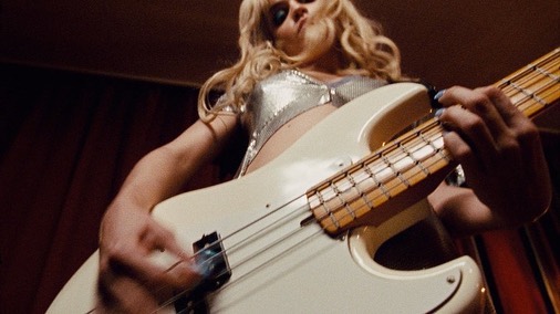 Low-angle shot of a blonde woman playing a white bass guitar indoors, wearing a shiny top, capturing a dynamic musical performance.