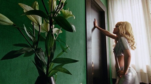 Woman with blonde hair poses by a dark door in a metallic outfit; a floral arrangement is in the foreground against a green wall.