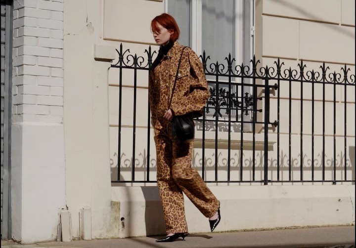 An adult woman in a leopard-print outfit and black accessories walks in an urban setting by a cream building and a black iron fence.
