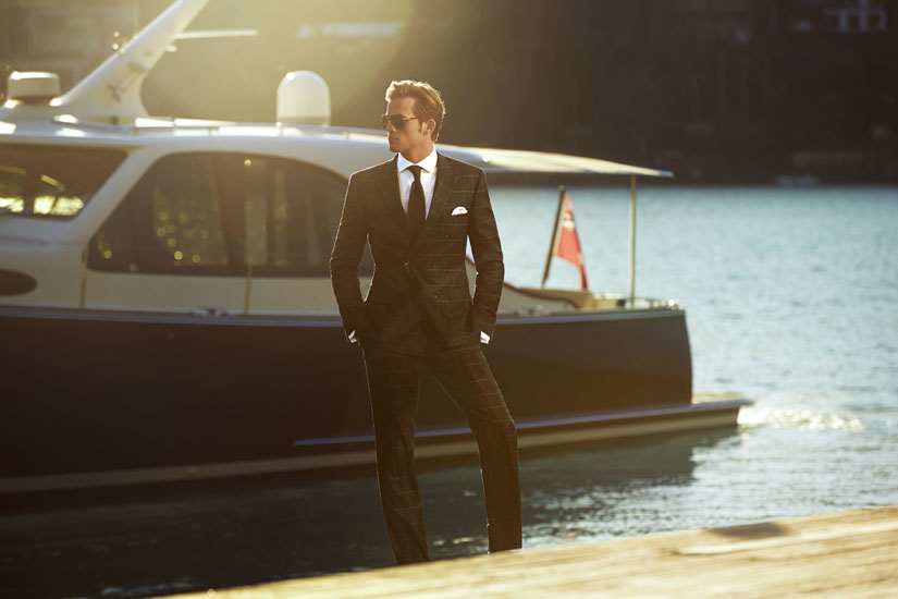 A man in a suit poses on a dock next to a yacht, wearing sunglasses in golden hour sunlight. View includes sea and coastal hills.