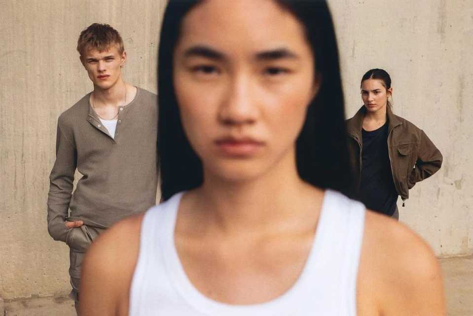 Fashion shot with three young adults in neutral clothing standing against a concrete wall with natural light in a medium shot.