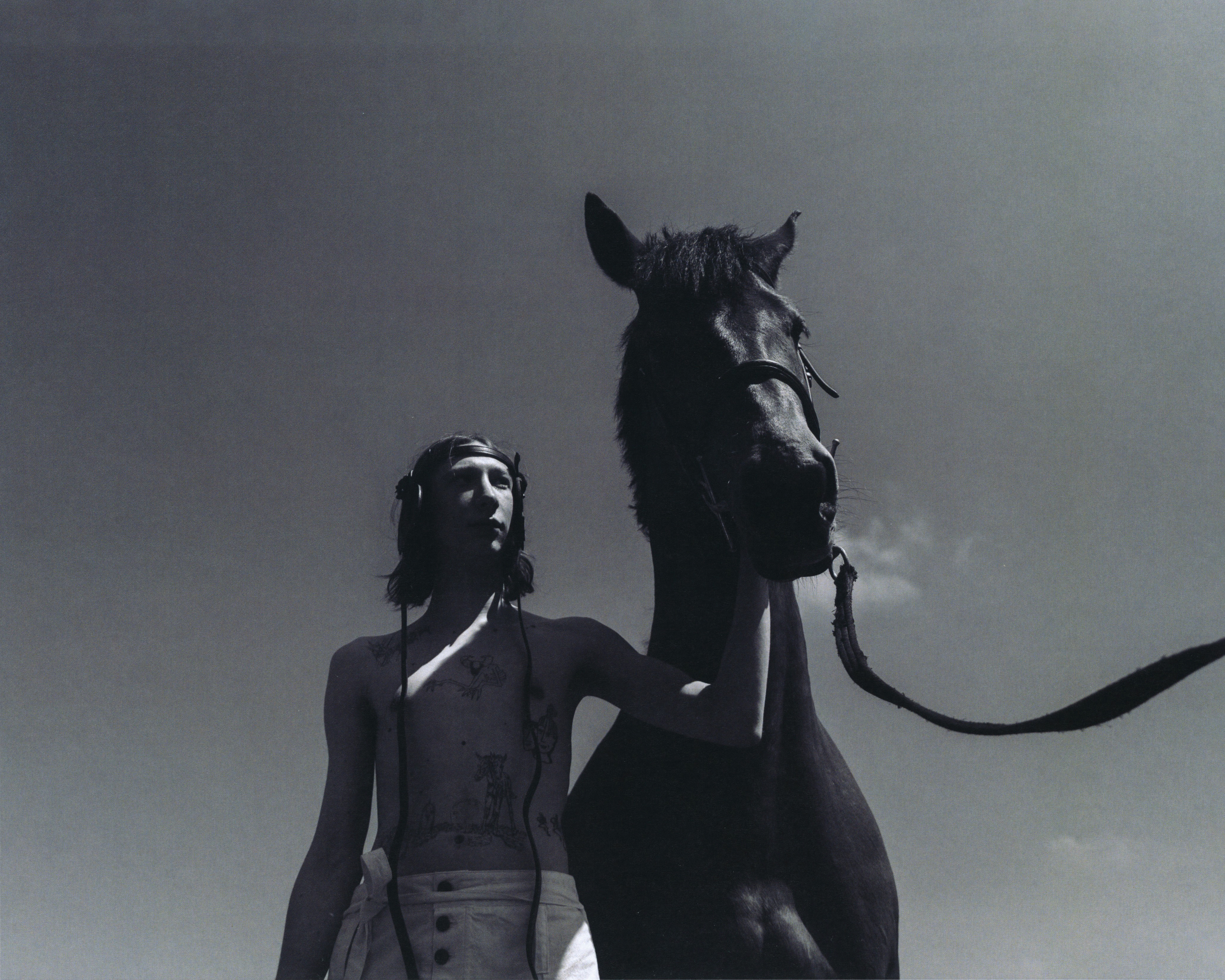 Monochromatic shot of a slender person standing next to a dark horse against a bright, cloudless sky, creating a striking silhouette.