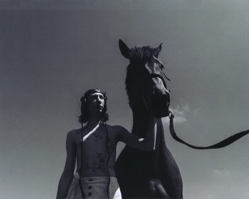 Monochromatic shot of a slender person standing next to a dark horse against a bright, cloudless sky, creating a striking silhouette.