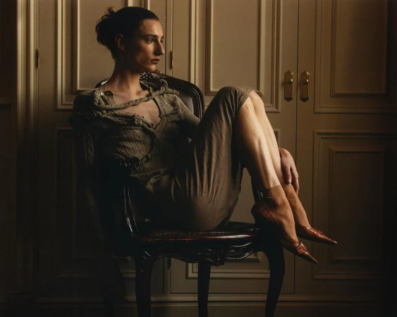 Model seated on a dark wooden chair with knees drawn up, wearing a textured knitted dress and pointed heels against panelled doors