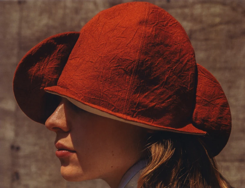 Profile view of a fair-skinned person wearing a large, textured, rusty-red hat with a beige brim, against a neutral, mottled background.