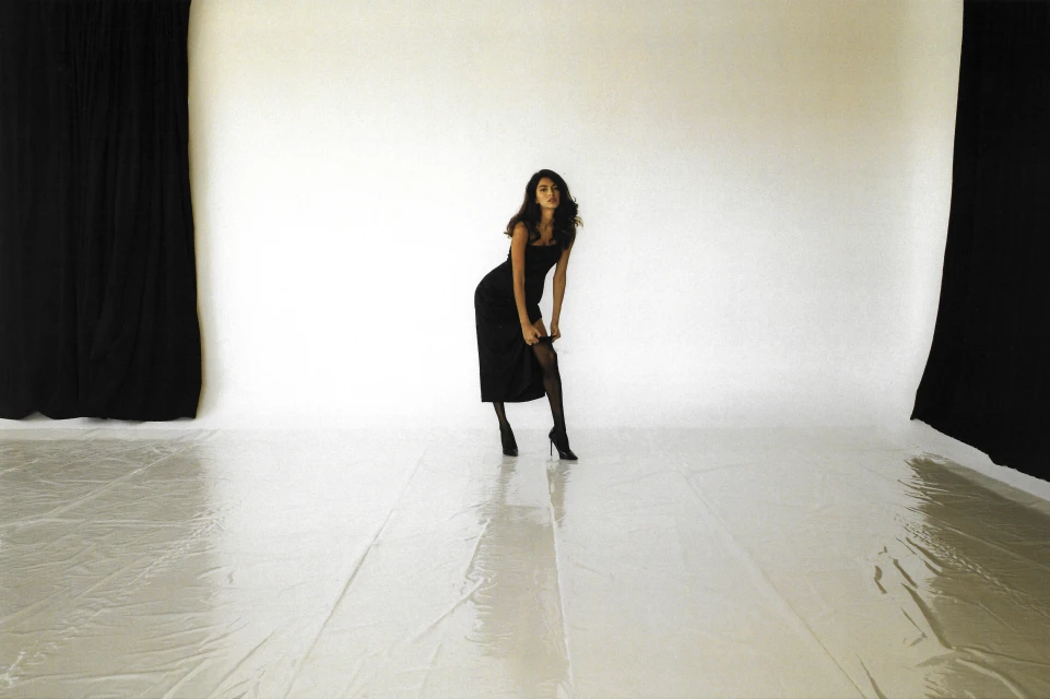 A slender woman in a black dress and heels poses in a photo studio with a white backdrop and black curtains, striking a confident pose.