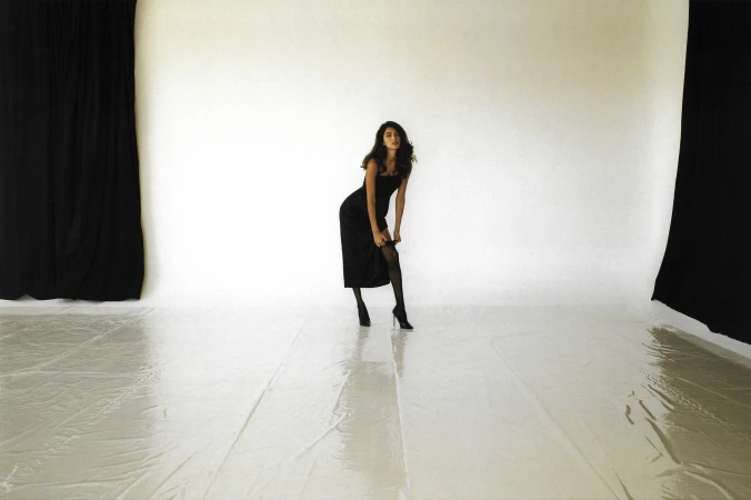 A slender woman in a black dress and heels poses in a photo studio with a white backdrop and black curtains, striking a confident pose.